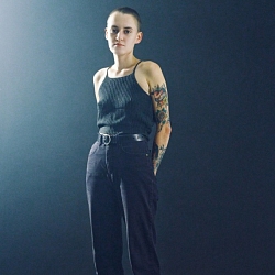 Photo of artist Sister Ray, in black pants and a black top, in front of a black background.