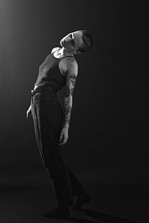 Photo of artist Sister Ray, in black pants and a black top, in front of a black background looking up. Photo Credit: Vanessa Heins