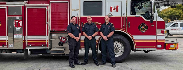 Image of three firefighters standing in front of a red and white fire truck with a ladder on top.