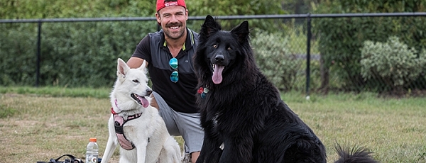 A kneeling man smiles with his two dogs, one white, one black, both of which are cute and fluffy.